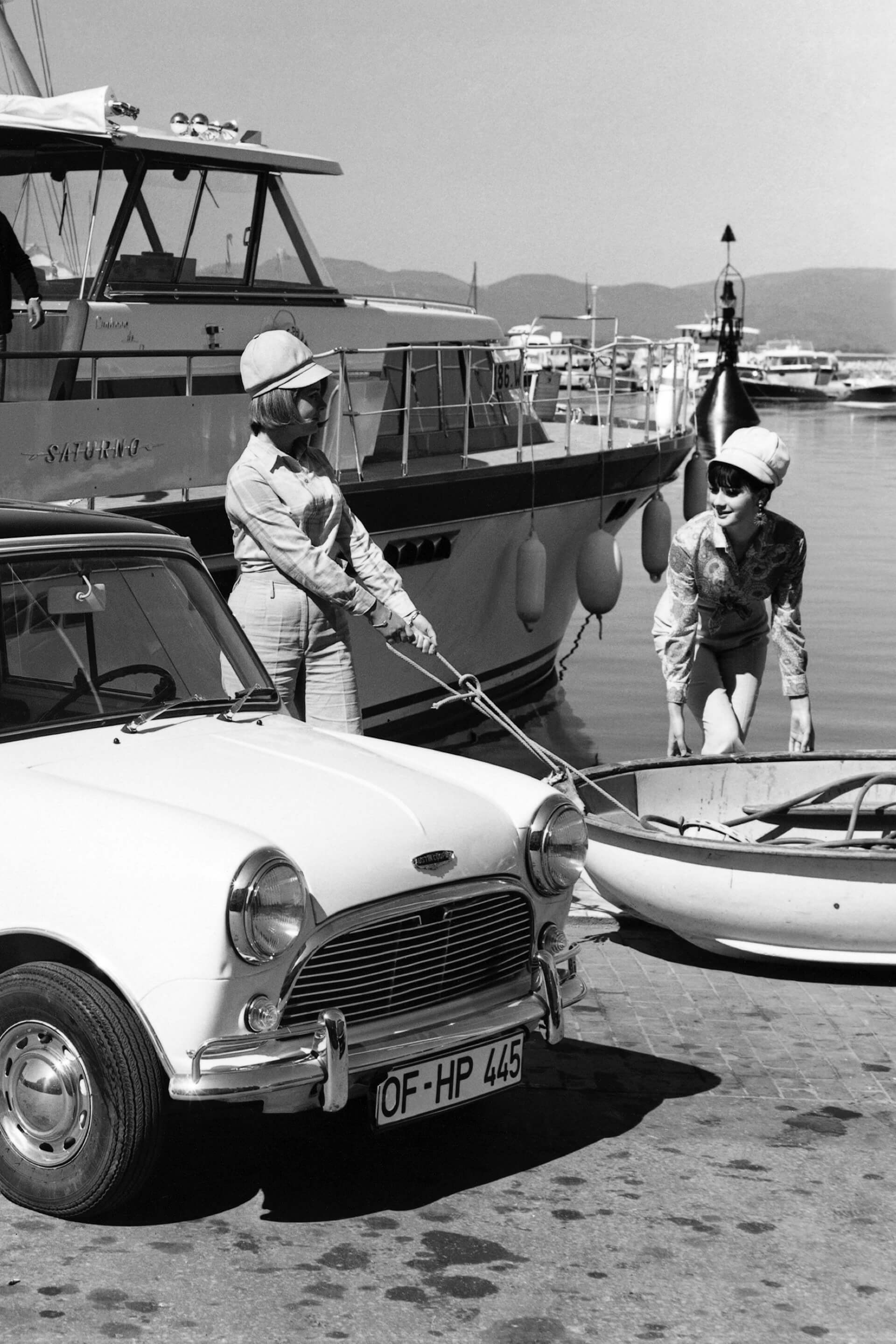 Morris Mini-Minor parked by the water's edge at a marina and two women prepare to board a dinghy nearby.